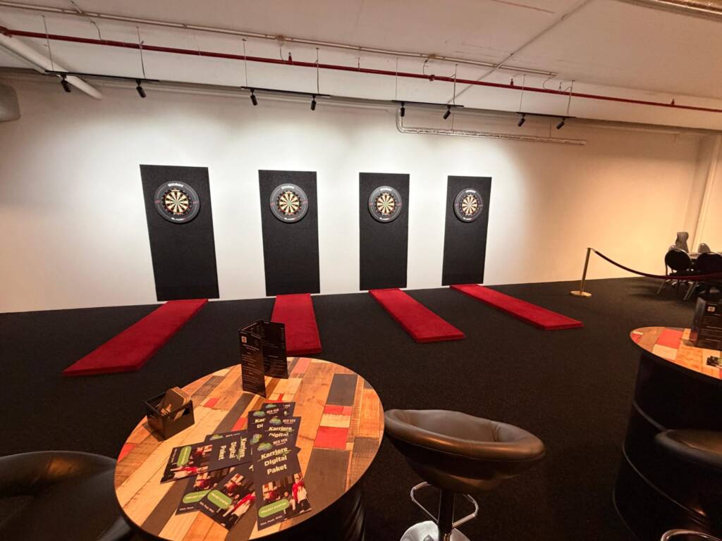 Ein moderner Dart-Raum mit vier beleuchteten Dartboards an einer weißen Wand. Davor liegen rote Wurfmatten. Im Vordergrund stehen hohe Tische aus schwarzen Fässern mit Barhockern. Die Beleuchtung ist warm und erzeugt eine entspannte Atmosphäre in Frontansicht.