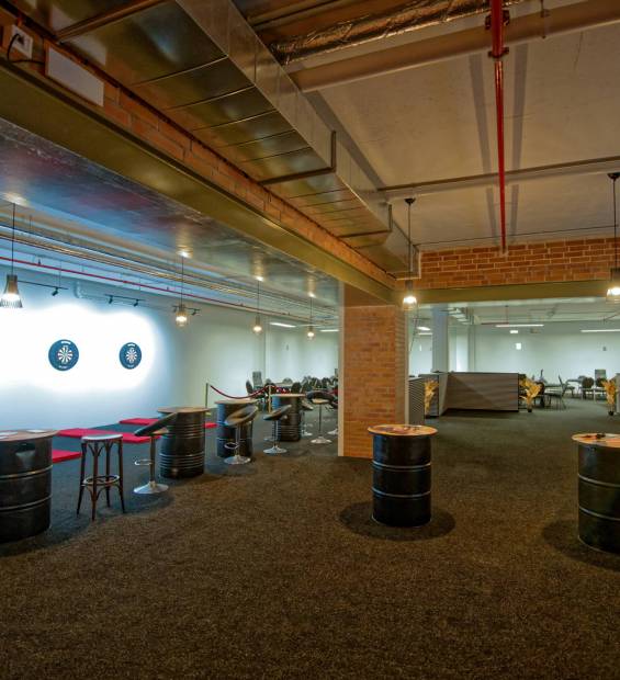 Ein großer Freizeitbereich mit vier beleuchteten Dartboards links, schwarzen Fass-Stehtischen im Raum und einem offenen Sitzbereich mit Tischen und Hängelampen im Hintergrund.