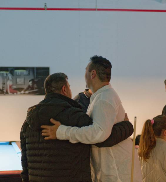 Mehrere Personen stehen in einem Billardraum beisammen, zwei Männer umarmen sich, ein Mädchen steht daneben.