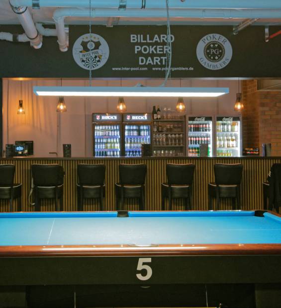 Ein Pooltisch mit der Nummer 5 steht im Vordergrund; dahinter eine beleuchtete Bar mit Getränkekühlschränken, Barhockern und einem Schild „Billard Poker Dart“.