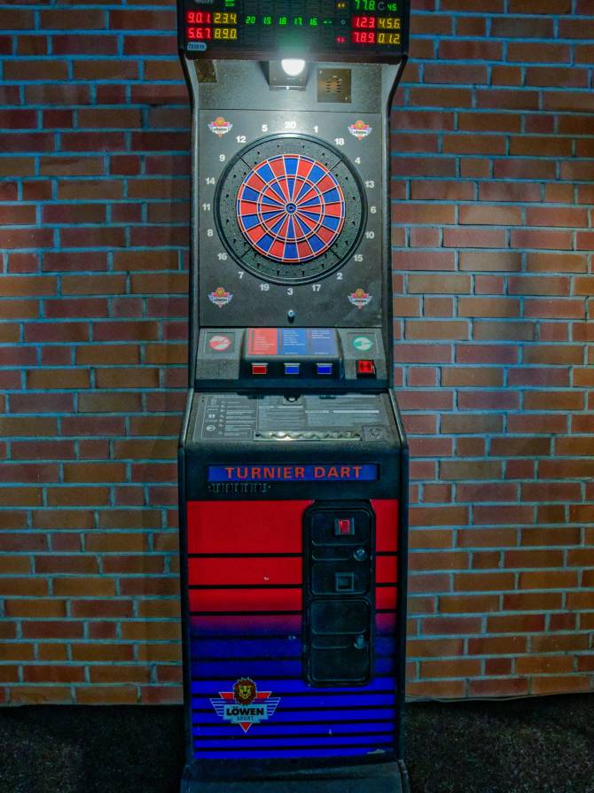 Ein elektronischer Turnier-Dartautomat vor einer Backsteinwand, mit beleuchteter Dartscheibe und digitalem Punktedisplay.