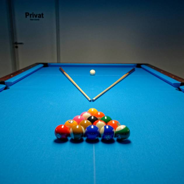 Ein Pooltisch mit aufgebautem Dreieck bunter Kugeln im Vordergrund, zwei Queues in V-Form dahinter und der weißen Kugel vor einer Tür mit der Aufschrift „Privat“.